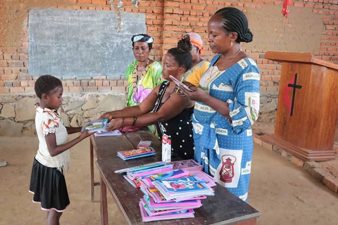 Younger children at the orphanage attend school. This young girl receives a book from the management team which includes Furaha Tshoso (right).  Photo by Chadrack Tambwe Londe, UM News.