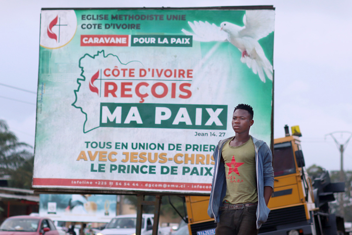 Un piéton passe devant l’un des panneaux publicitaires de l’Église Méthodiste Unie appelant à la paix dans la commune d’Abobo, en Côte d’Ivoire. L’Église Méthodiste Unie et ses partenaires ont organisé une campagne de communication demandant à la population et aux autorités de faire preuve de retenue pendant la période électorale. La caravane a traversé plusieurs villes et hameaux de Côte d’Ivoire. Photo d’Isaac Broune, UM News.