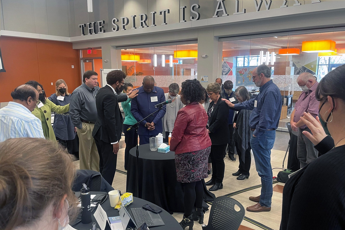 Miembros de la Mesa Conexional imponen las manos sobre el Obispo Mande Muyombo presidente de la Mesa Conexional, al centro, y Judi Kenaston directora interina de ministerios conexionales, cuarta de derecha a izquierda, durante la reunión del grupo el 24 de febrero en Atlanta, Georgia. El cuerpo de liderazgo ayuda a determinar el presupuesto de La Iglesia Metodista Unida. 