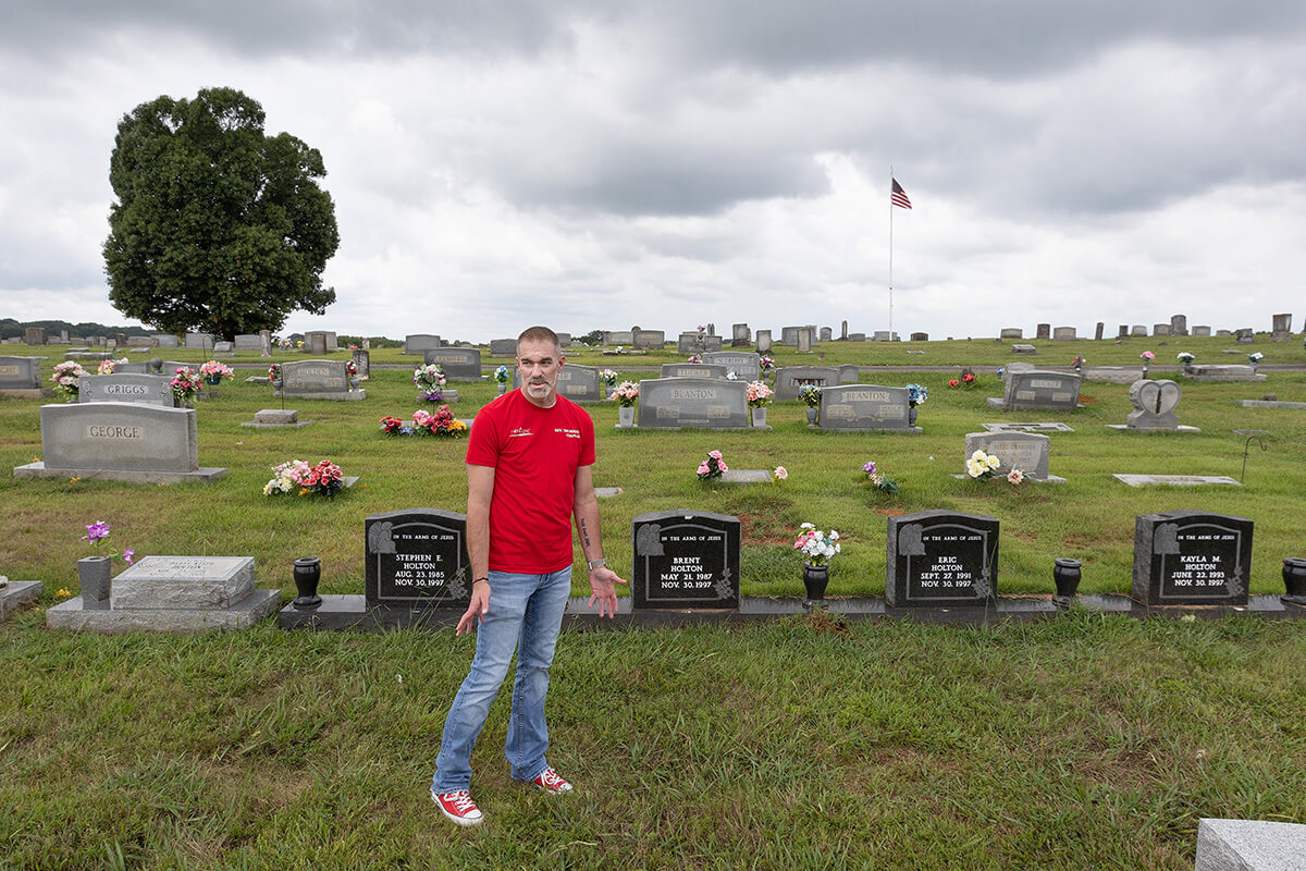 O Rev. Tim Holton, um pastor Metodista Unido, visita túmulos de família no Cemitério Simpson em Eagleville, Tennessee. Em 1997, seu primo, Daryl Holton, matou seus quatro filhos com um rifle de estilo militar e acabou sendo executado na cadeira elétrica do Tennessee. Ele está enterrado sob a lápide de cor clara à esquerda, ao lado dos túmulos de seus filhos. Tim Holton agora serve no conselho do Tennesseans por Alternativas à Pena de Morte e ministra a presos condenados à morte como capelão voluntário na Instituição de Segurança Máxima Riverbend em Nashville, Tennessee. Foto de Mike DuBose, Notícias MU.