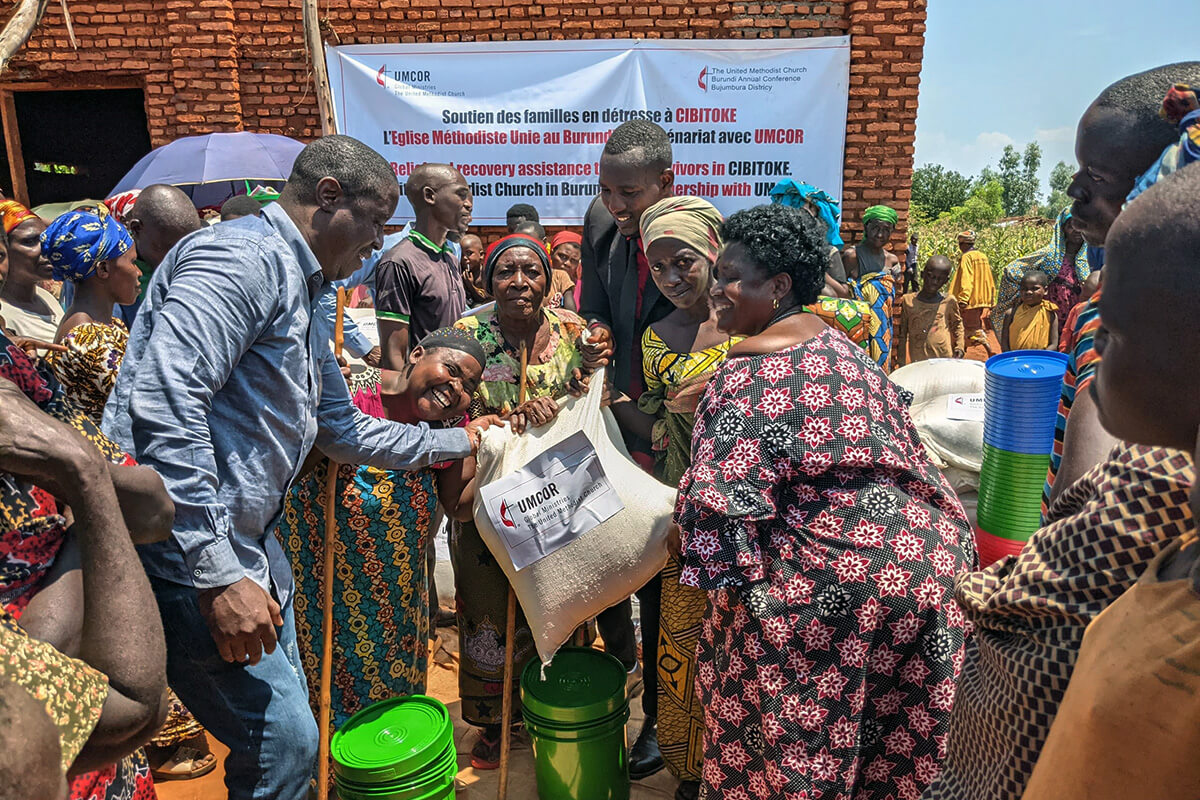 Patrick Abro (à gauche), missionnaire Méthodiste Uni occupant le poste de responsable des opérations sanitaires au sein de la Conférence du Burundi, et le révérend Cimpaye Valentine (à droite), surintendant du district de Bujumbura, remettent un sac de riz à des survivants des inondations à Cibitoke, au Burundi. Grâce au soutien financier du Comité Méthodiste Uni pour le Secours, l'Église a aidé 140 foyers touchés par les graves inondations qui ont frappé le district. Photo de Jérôme Ndayisenga, UM News.